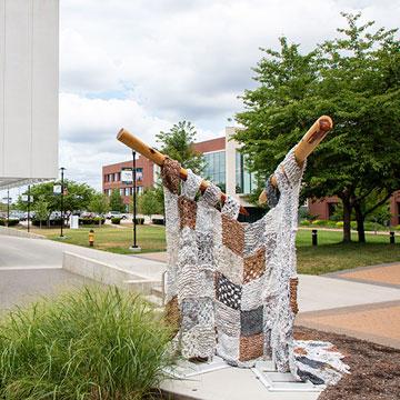 “One Stitch at a Time” by art student Alayna Reinke. The sculpture is a giant knitted afghan on giant knitting needles. The "yarn" is plastic bags.